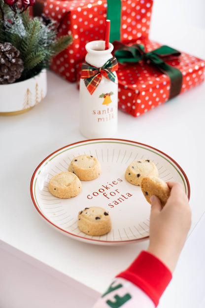 Cookies on a plate with 'Cookies for Santa' text, next to a bottle labeled 'Santa Milk', with Christmas presents and decorations in the background.