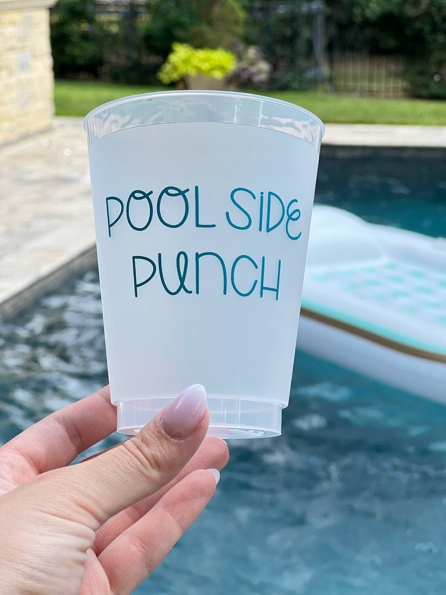 Clear plastic cup with 'Pool Side Punch' text held by a hand by a pool.